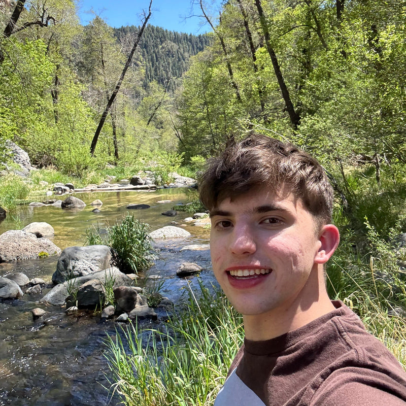 Person taking a selfie by a river with trees and rocks in the background